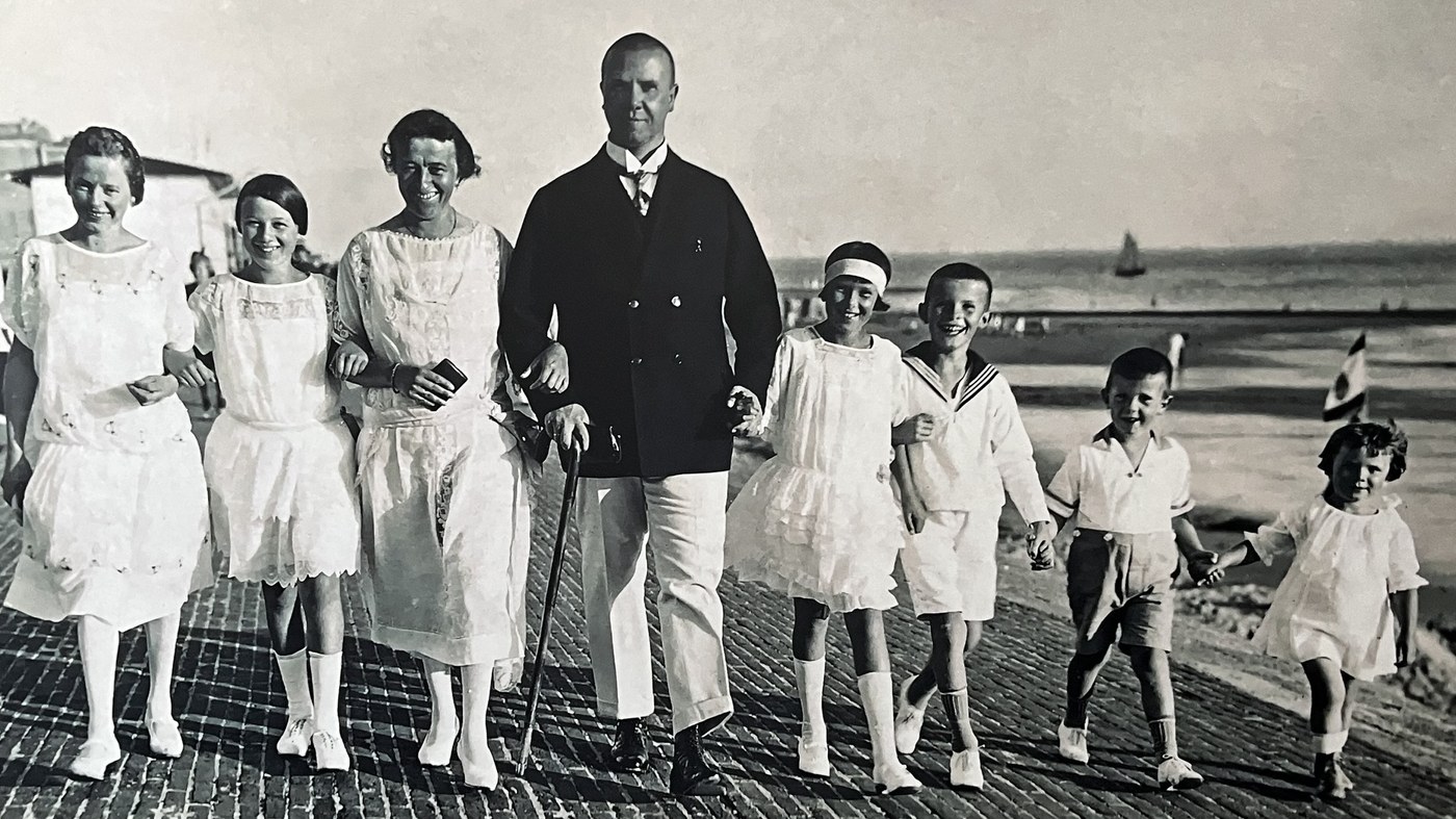 Familie Joseph Schilgen in einer Aufnahme auf Norderney: Joseph mit Anzug, Stock und Hut, daneben drei Frauen und vier Kinder in heller Sommerkleidung, im Hintergrund Strand und Segelboot